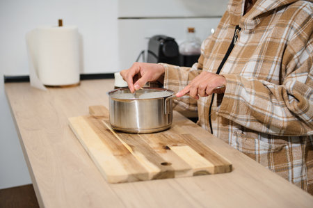 A woman in a plaid hoodie stirs a saucepan on a wooden board in a modern kitchen. The scene exudes...
