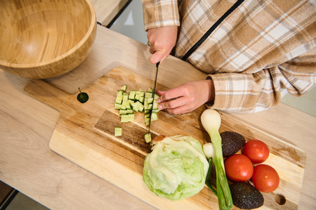 A person chops fresh vegetables, including cucumbers and lettuce, for a nutritious homemade salad. The setup includes kitchen tools, showcasing healthy food preparation in a cozy and inviting setting.の写真素材