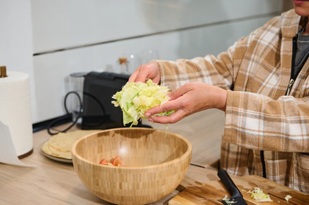 A person prepares a healthy salad in a wooden bowl, focusing on fresh greens and vegetables. The setting is a home kitchen, promoting healthy eating practices in a comfortable environment.の写真素材