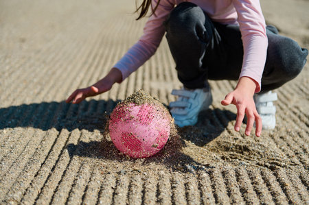 Child kneeling on a striped sandy beach, lifting a pink ball partially covered with sand, suggesting playfulness and outdoor fun.の写真素材
