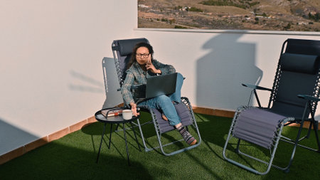 A woman working remotely on a laptop with a phone call on a sunny outdoor space.の写真素材
