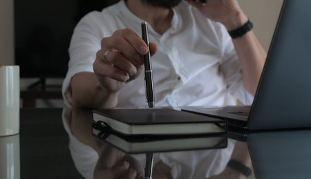 Man in a modern workspace using a laptop and notebook for work, demonstrating focus and productivity. Represents freelance or remote work lifestyle.の写真素材