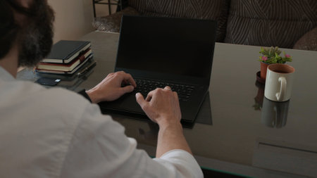A man in a professional workspace uses a laptop computer while surrounded by notebooks, a plant, and...