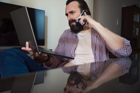 Focused young man managing tasks on a laptop while talking over a smartphone in home office.