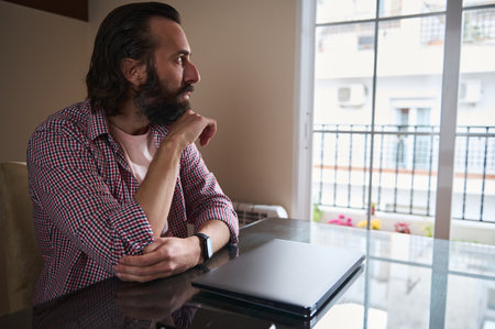 A contemplative man sits by a laptop, gazing through a large window, conveying focus and introspection in a personal workspace setting.の写真素材