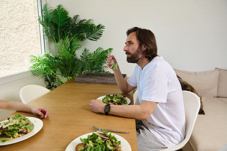 A man sits at a wooden table beside a friend, enjoying a fresh salad meal in a bright, homey...