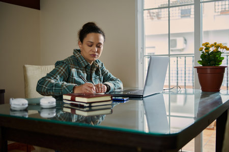 A concentrated woman working at her desk, taking notes on a journal next to her laptop. The...