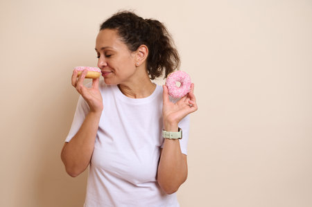 A cheerful woman cherishes two pink frosted doughnuts, savoring their scent. Representing indulgence and happiness, this scene invokes happiness and culinary delight.の写真素材