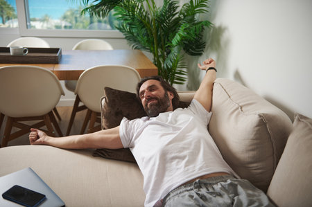 A relaxed man stretches comfortably on a sofa in a modern living room with natural decor and bright daylight enhancing the tranquil atmosphere.の写真素材