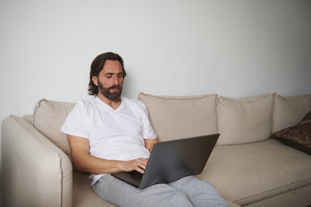 A man in casual attire typing on a laptop while seated on a beige sofa in a home environment, reflecting relaxation and productivity.の写真素材