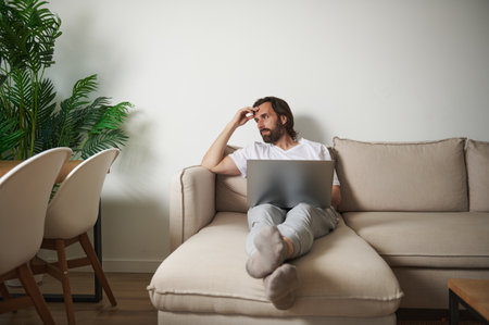 Professional man working remotely on a laptop in a cozy living room space, reflecting on ideas....