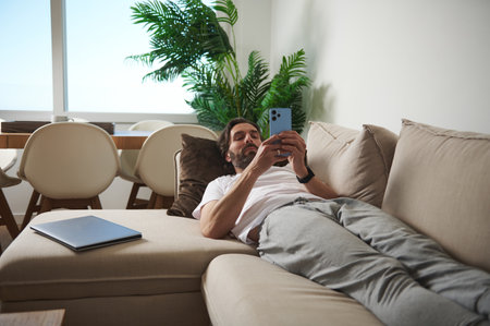 A man reclining on a sofa uses his smartphone in a bright, minimalist living room with natural...