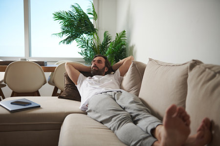 Man resting reclined on a soft sofa in a bright and tranquil living room. The space exudes comfort and simplicity, providing an atmosphere perfect for relaxation.の写真素材