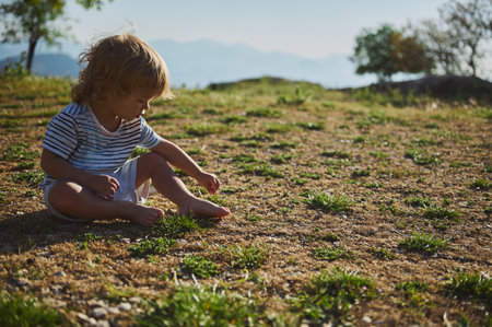 A young child sits relaxed on a sunny day, playing and exploring a grassy area. The outdoor atmosphere emphasizes peace, curiosity, and connection with nature.の写真素材