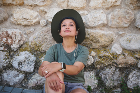 A woman wearing a green shirt and hat sits relaxed against an aged stone wall, reflecting serenity...