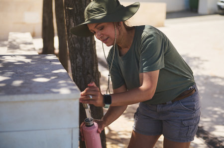 A woman refills her bottle at an outdoor fountain beneath summer sunlight. She wears a wide-brimmed...