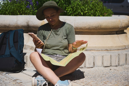 Traveler seated outdoors holding a smartphone and examining a map, enjoying a sunny day while exploring a historic city. A blue backpack and stonework create a relaxed, adventurous atmosphere.の写真素材