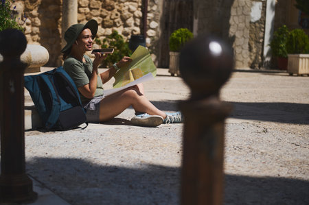 A tourist sits in a historical town square, holding a map and using mobile navigation. Surrounded by traditional architecture, they exude curiosity and adventure.の写真素材
