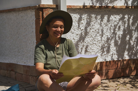 A cheerful traveler enjoys reading a map outdoors in a sunny area, wearing a comfortable hat and...