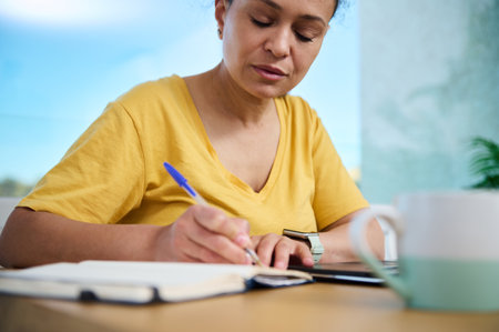A woman sits at a desk in a bright home interior, writing in a notebook and working with a tablet, symbolizing productivity, planning, and focus within a flexible work environment.の写真素材