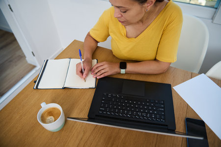 A woman writes notes in a notebook at home, accompanied by a laptop and a cup of coffee, depicting...