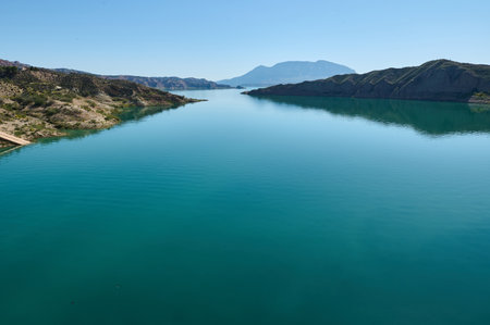 Beautiful scene of a serene lake with reflective blue water, surrounded by arid hills and overlooked by a distant mountain under a cloudless sky.の写真素材