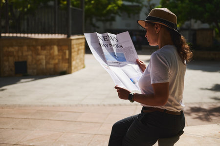 A woman sits outdoors in the sunlight, reading a newspaper. She wears a hat and casual attire, creating a peaceful and relaxed scene amidst warm ambient lighting.の写真素材