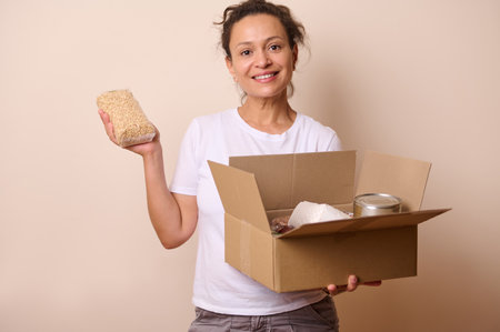 A cheerful woman holding a cardboard box with food items and a packet of grains. Represents...