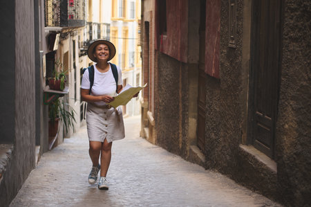 Tourist woman smiling while traversing a charming old town. With a map, she explores narrow historic streets on a sunny day, enjoying the journey and discovery.の写真素材