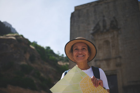 A cheerful woman traveler holds a map while exploring a picturesque historic city. She enjoys discovering ancient architecture under the warm sunlight, embodying the joy of travel and exploration.の写真素材