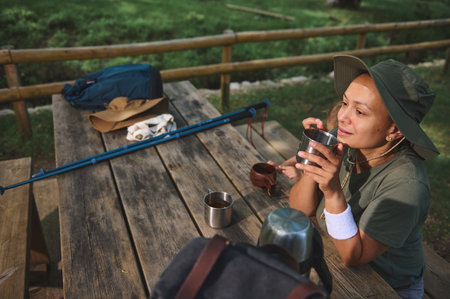 A woman relaxes at a wooden outdoor table, enjoying a drink in a peaceful natural environment, highlighting moments of leisure and appreciation for outdoor lifestyles.の写真素材