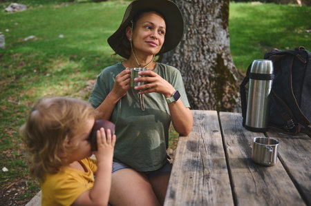 A woman and her child share a peaceful moment outside, drinking beverages. They sit on a wooden bench under green trees, embracing nature and family togetherness.の写真素材