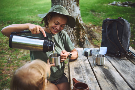 A smiling woman sits outdoors, engaging in a relaxed moment, pouring a drink for her child on a wooden picnic table surrounded by greenery. Features a natural and inviting setting.の写真素材