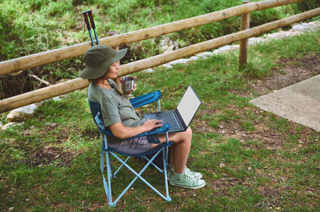 A woman enjoys outdoor work life, sitting in a folding chair amidst nature. Equipped with a laptop...