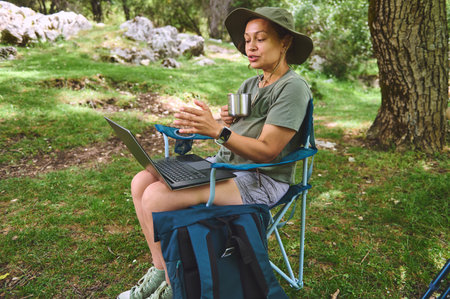 A woman sits in a camping chair outdoors, working on a laptop while holding a coffee mug amidst lush...