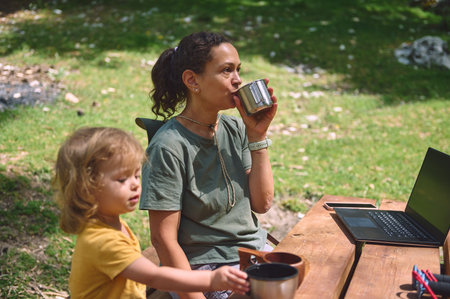 A mother enjoys the outdoors while working remotely on her laptop, drinking from a mug, with her child nearby. The scene depicts work-life balance in a natural setting.の写真素材