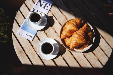 Image of a wooden table with cups of coffee, crispy croissants, and a newspaper, creating a tranquil...