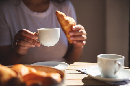 A close-up view of a woman holding a coffee cup and croissant, enjoying a peaceful morning breakfast under warm sunlight. Coffee and relaxation in a serene atmosphere.の写真素材