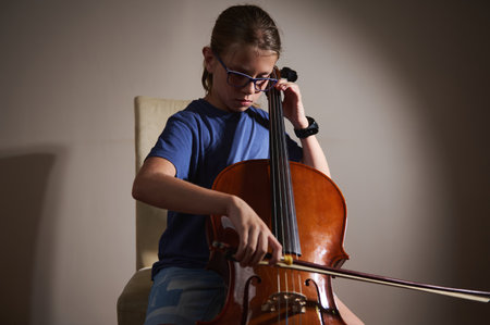 A young musician wearing glasses plays the cello with concentration in a minimalistic setting, showcasing dedication and musical talent.の写真素材