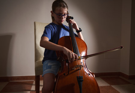 A child musician practices the cello in a quiet setting, sitting on a chair with focused concentration. The warm lighting highlights her dedication to mastering the instrument.の写真素材