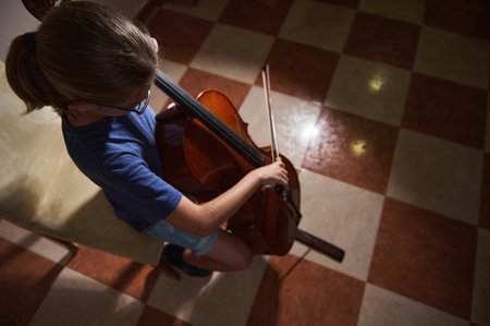 A young individual practices cello, seated on a chair in a well-lit room with a patterned floor, symbolizing focus and passion for music.の写真素材