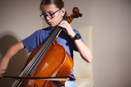 A young girl sits focused as she plays the cello in a warmly lit room, embodying creativity and passion for music.の写真素材