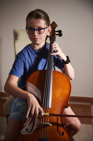 A young musician plays the cello with concentration and skill, seated in a natural, comfortable setting. The warm lighting highlights the instrument and focused determination of the performer.の写真素材