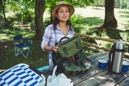 Woman organizing her backpack at a wooden table during a picnic, surrounded by greenery and...