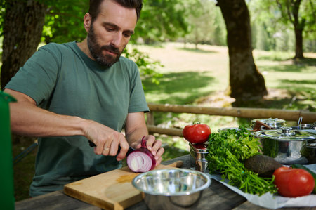 A man chops vegetables on a wooden table surrounded by fresh ingredients. The serene outdoor...