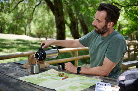 A man enjoys the outdoors while pouring a drink from a thermos near maps and equipment, embracing...