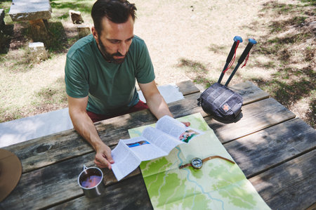 A man sits at a wooden table in a park, studying a map and compass, surrounded by trekking gear and enjoying nature.の写真素材