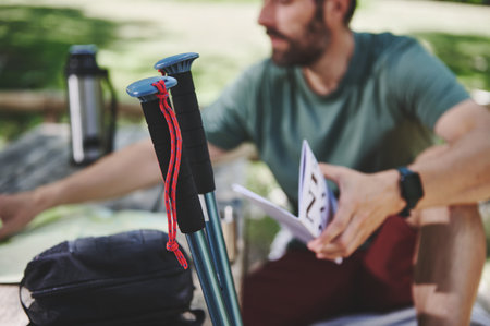 Trekking poles leaned against a backpack on a wooden surface with a person exploring navigational items. A serene outdoor setting under the sun.の写真素材