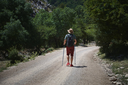 A man walks along a dirt path surrounded by greenery, wearing a backpack and hat, holding trekking poles, on an outdoor exploration adventure.の写真素材