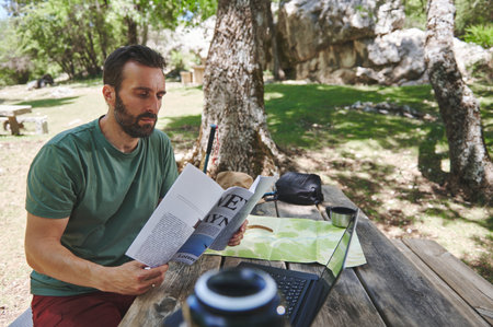 A man enjoying a tranquil moment reading outdoors at a picnic table, surrounded by nature. A laptop, a mug, and a map sit on the table, emphasizing exploration.の写真素材